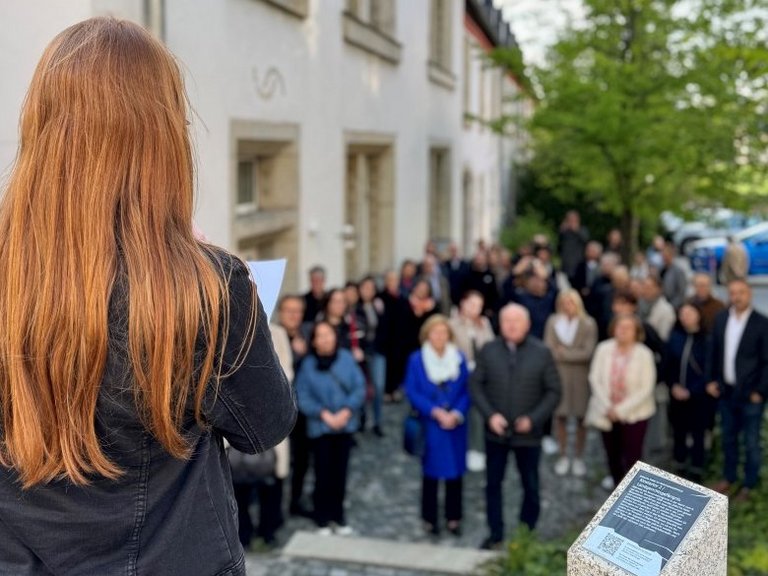 Auf dem Foto ist eine Frau von hinten zu sehen, die zu einer Menschenmenge spricht. Neben ihr steht eine Stele mit Infotext.