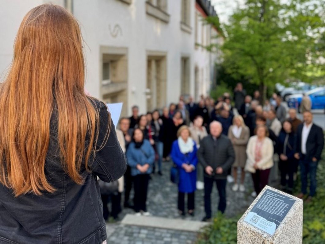 Auf dem Foto ist eine Frau von hinten zu sehen, die zu einer Menschenmenge spricht. Neben ihr steht eine Stele mit Infotext.