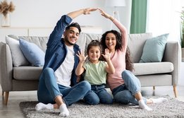Auf dem Bild ist ein Paar zu sehen, das in einem Wohnzimmer die Arme über ein kleines Mädchen hält.