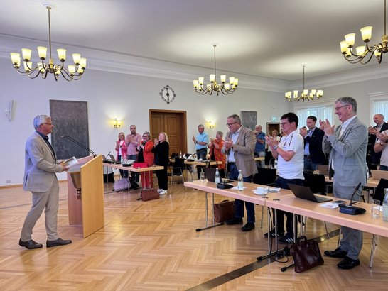 Auf dem Bild ist ein Mann an einem Pult zu sehen, dem ein Publikum im Stehen applaudiert.