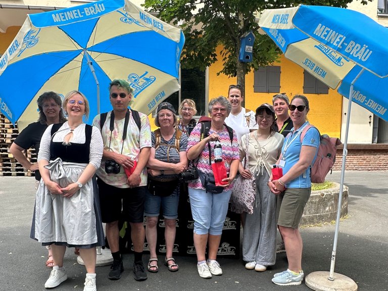Auf dem Bild sind zehn Personen unter Sonnenschirmen mit der Aufschrift Meinel-Bräu zu sehen.