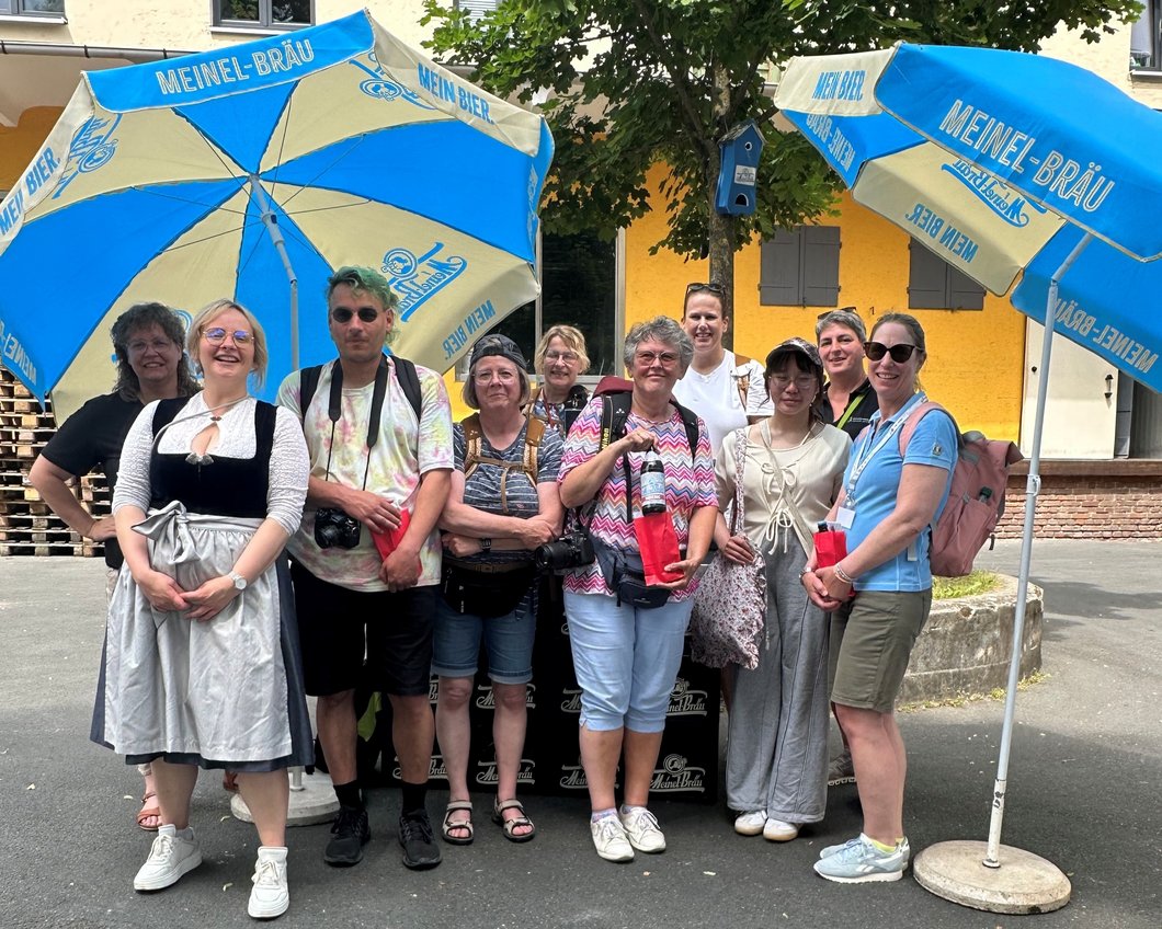 Auf dem Bild sind zehn Personen unter Sonnenschirmen mit der Aufschrift Meinel-Bräu zu sehen.