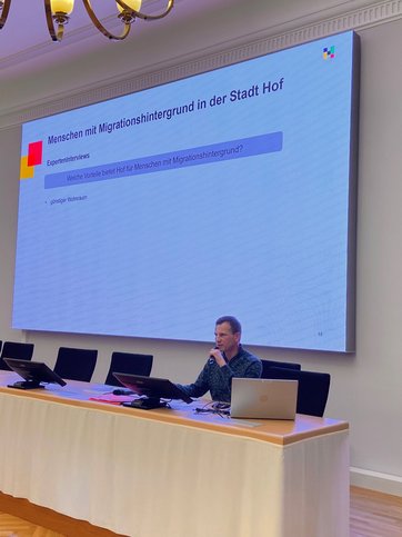 Auf dem Bild ist ein Referent zu sehen, hinter ihm eine Präsentation über Menschen mit Migrationshintergrund in Hof.
