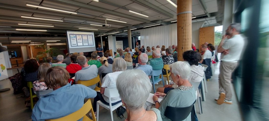 Auf dem Bild sind zahlreiche Menschen zu sehen, die einem Vortrag mit Powerpoint-Präsentation lauschen.