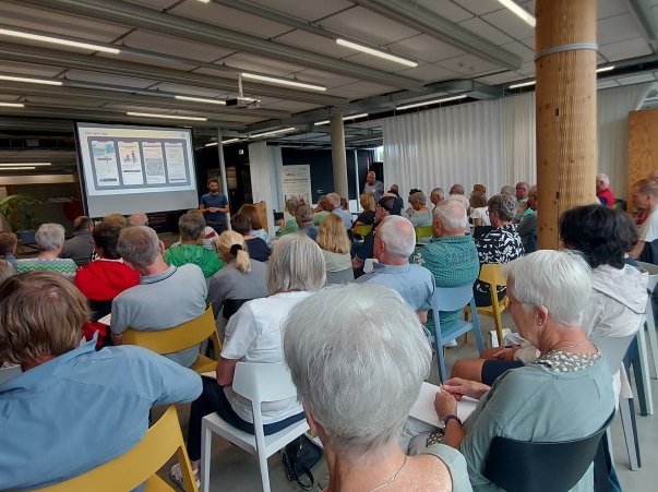 Auf dem Bild sind zahlreiche Menschen zu sehen, die einem Vortrag mit Powerpoint-Präsentation lauschen.
