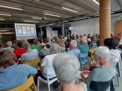 Auf dem Bild sind zahlreiche Menschen zu sehen, die einem Vortrag mit Powerpoint-Präsentation lauschen.