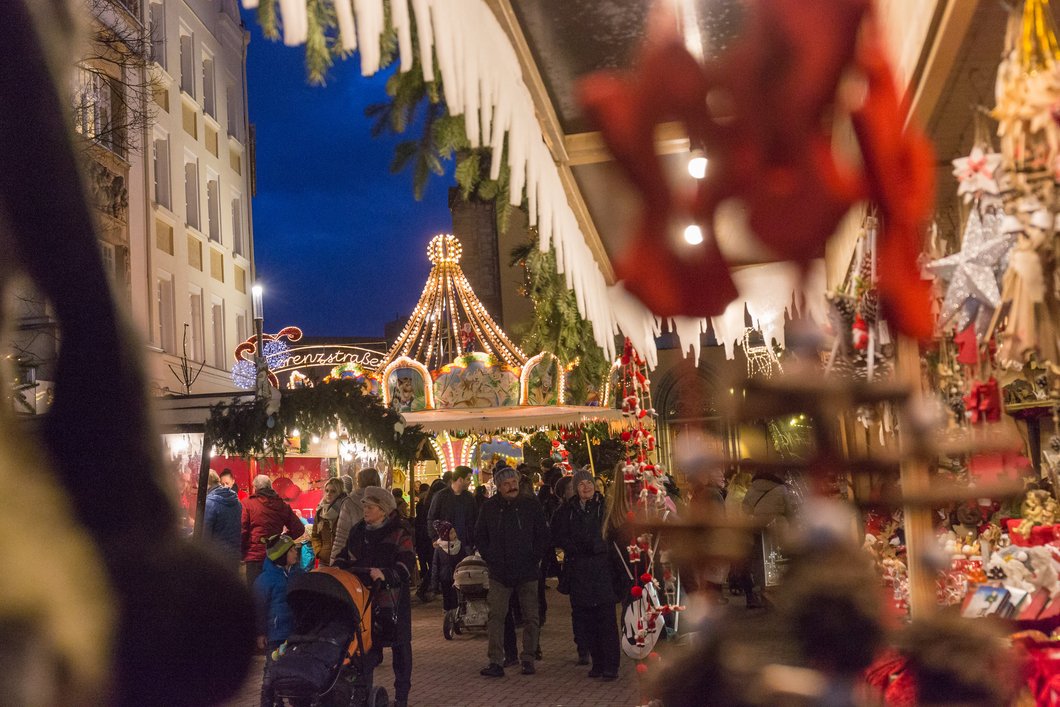 Auf dem Bild sind Menschen zu sehen, die abends über den hell erleuchteten Hofer Weihnachtsmarkt schlendern.