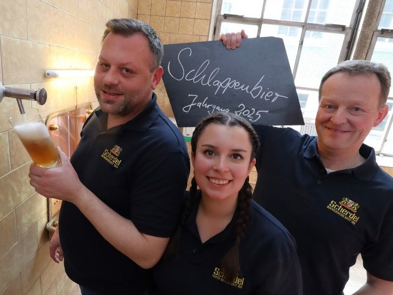 Auf dem Foto sind zwei Männer und eine Frau in einer Brauerei zu sehen. Ein Mann nimmt eine Sudprobe aus einem Hahn, der andere hält ein Schild mit der Aufschrift "Schlapplenbier Jahrgang 2025" hoch.