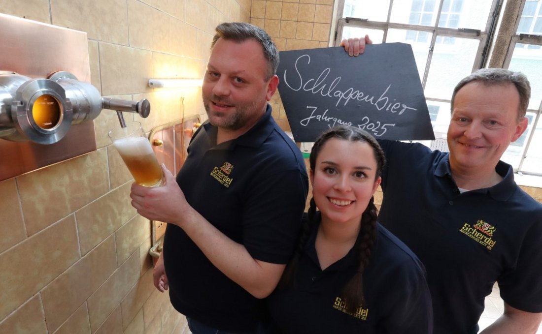 Auf dem Foto sind zwei Männer und eine Frau in einer Brauerei zu sehen. Ein Mann nimmt eine Sudprobe aus einem Hahn, der andere hält ein Schild mit der Aufschrift "Schlapplenbier Jahrgang 2025" hoch.