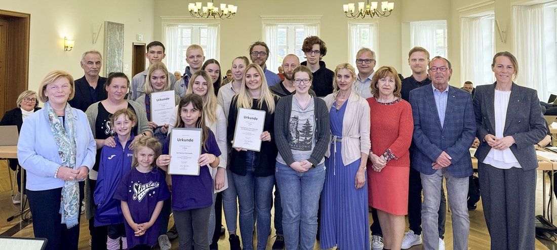 Auf dem Gruppenbild sind über 20 Personen zu sehen, die in einem Saal stehen. Zwei junge Frauen und ein Mädchen halten Urkunden hoch.