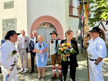 Auf dem Bild sind mehrere Personen zu sehen, die draußen stehen. Eine Frau hält einen Blumenstrauß.