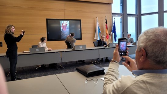 Auf dem Bild sind sechs Personen in einem Sitzungssaal zu sehen. Auf einem Monitor an der Wand wird ein Inhalt gezeigt, den sich drei der Personen gleichzeitig auf Handys ansehen.