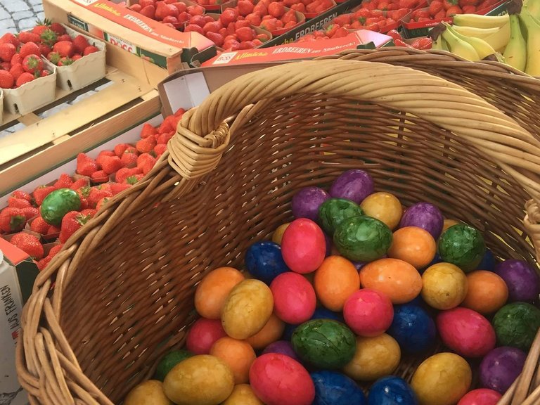 Auf dem Bild ist ein Korb mit bunt gefärbten Ostereiern zu sehen, dahinter Kisten mit Erdbeeren.