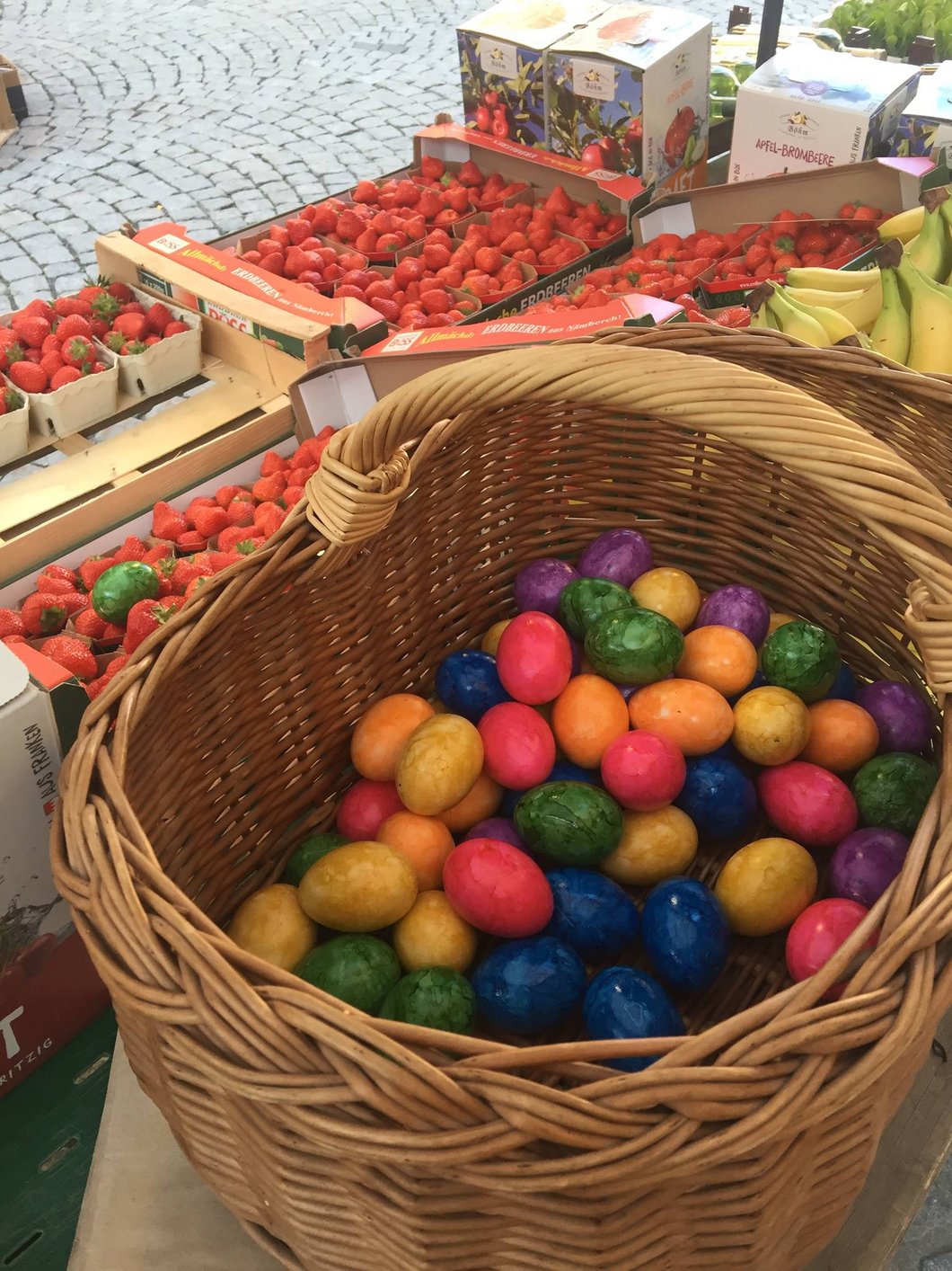 Auf dem Bild ist ein Korb mit bunt gefärbten Ostereiern zu sehen, dahinter Kisten mit Erdbeeren.