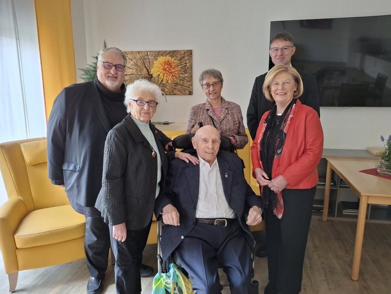 Auf dem Bild sind sechs Personen in einem Gemeinschaftsraum zu sehen. In der Mitte sitzt ein alter Mann im Rollstuhl, eine alte Frau legt ihm die Hand auf die Schulter.