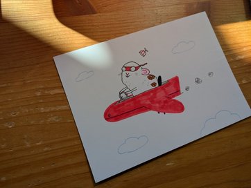 Auf dem Bild ist ein Blatt Papier mit einer Zeichnung zu sehen, die einen maskierten Hamster zeigt, der ein Flugzeug fliegt.