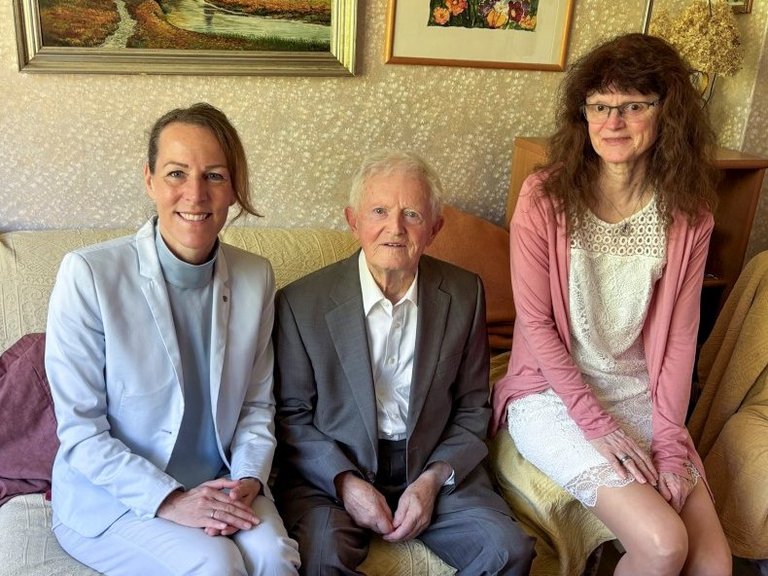 Auf dem Bild ist ein alter Mann zu sehen, der mit zwei Frauen in einem Wohnzimmer sitzt.