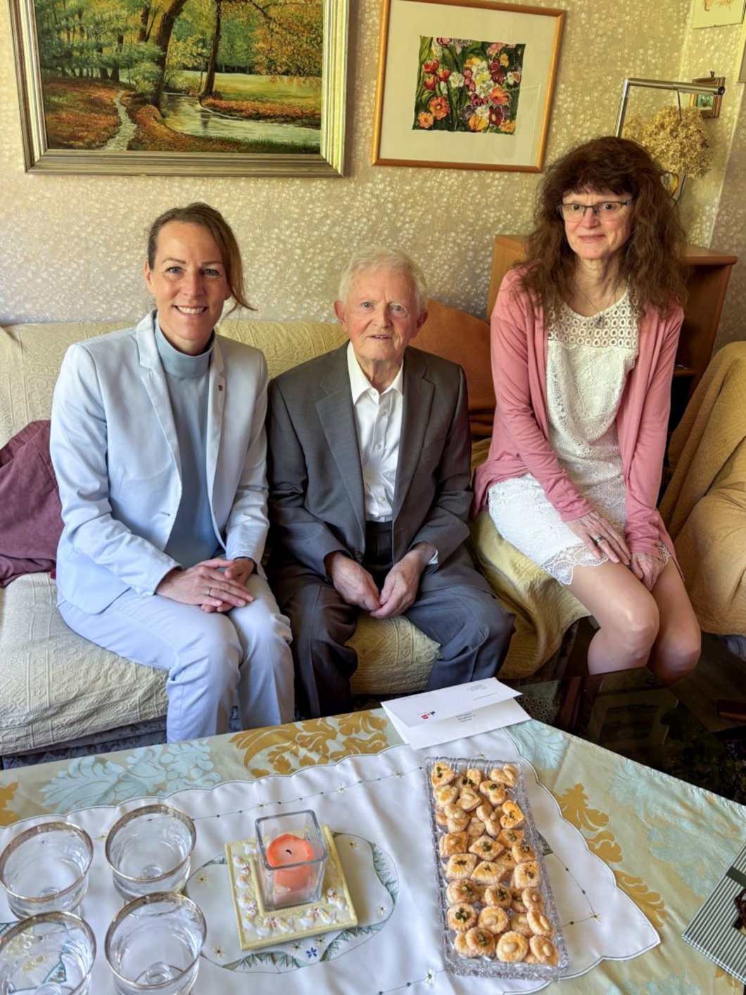 Auf dem Bild ist ein alter Mann zu sehen, der mit zwei Frauen in einem Wohnzimmer sitzt.