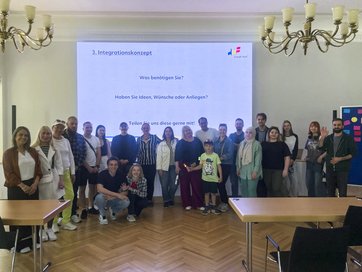 Über 20 Personen stehen in einem Saal vor einer Videopräsentation mit dem Titel Integrationskonzept.