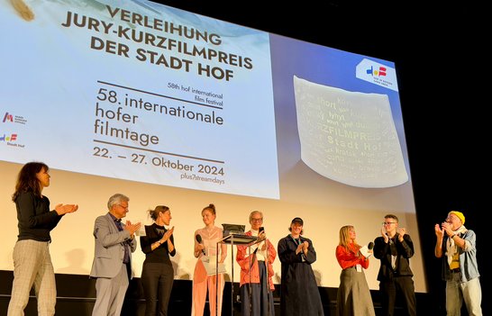 Auf dem Bild sind neun Personen zu sehen, die auf vor einer Kinoleinwand stehen. Eine junge Frau hält einen Preis in der Hand, die anderen klatschen.