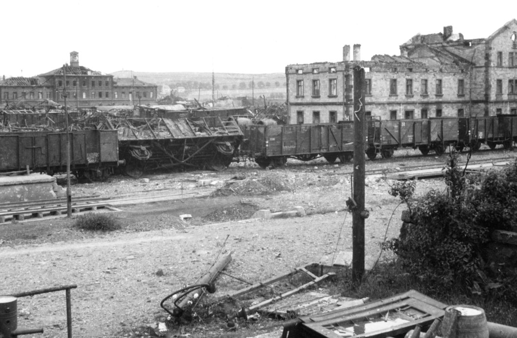 Auf dem Schwarz-Weiß-Foto sind Zugwagen zu sehen, die teils umgefallen und beschädigt sind. Im Hintergrund zerbombte Bahnhofsgebäude.