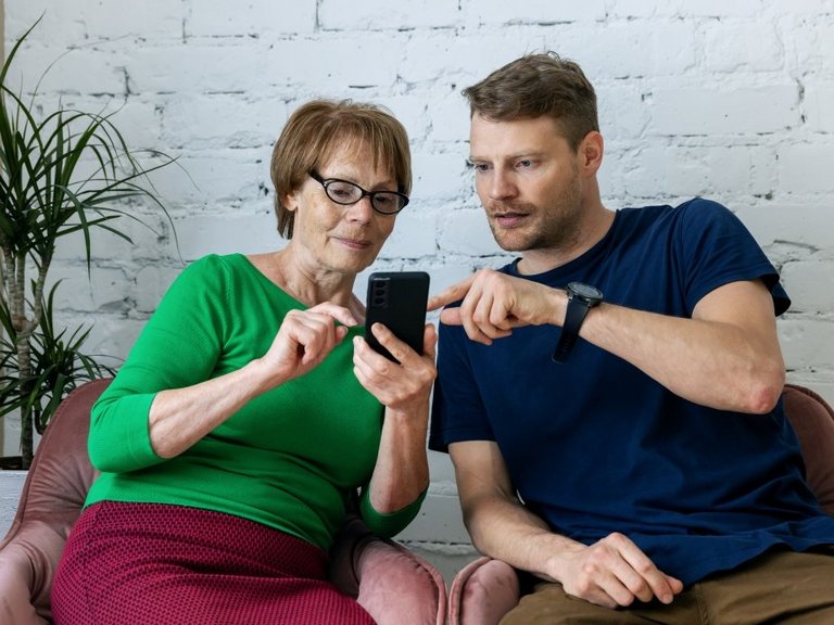 Auf dem Bild ist ein junger Mann zu sehen, der einer älteren Frau etwas an ihrem Smartphone erklärt.