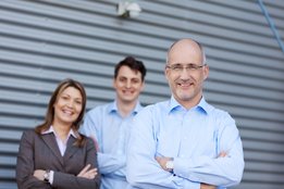 Auf dem Bild ist ein älterer Mann mit Brille zu sehen, hinter ihm stehen eine junge Frau und ein junger Mann, alle in Businesskleidung. 