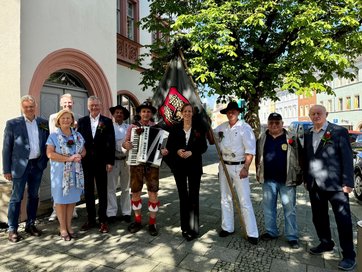 Auf dem Bild sind zehn Personen zu sehen. Zwei von ihnen tragen traditionelle Handwerkerkleidung und eine Fahne. Ein dritter Mann hält ein Akkordeon.
