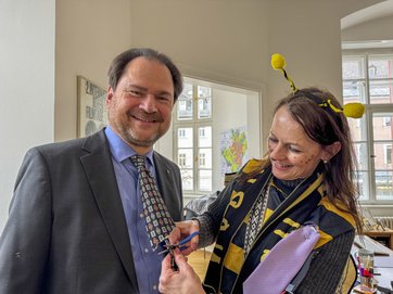 Auf dem Bild ist eine Frau im Bienenkostüm zu sehen, die einem Mann die Krawatte abschneidet.